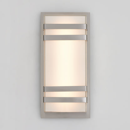 A modern rectangular wall sconce with a frosted glass panel, framed by a metallic border and three horizontal metal bars across the front, emitting soft, diffused light against a plain light gray wall.