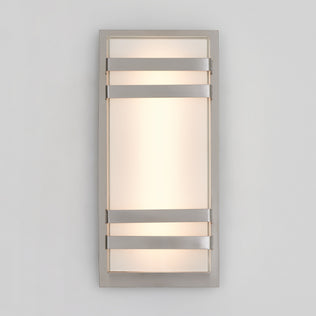 The Artika Glowbox 3 indoor-outdoor LED wall light features a frosted glass center with a metallic frame and three horizontal metal bands, casting soft, diffused light onto a light gray wall.