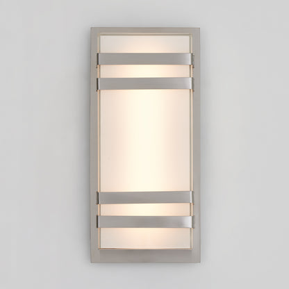 The Artika Glowbox 3 indoor-outdoor LED wall light features a frosted glass center with a metallic frame and three horizontal metal bands, casting soft, diffused light onto a light gray wall.