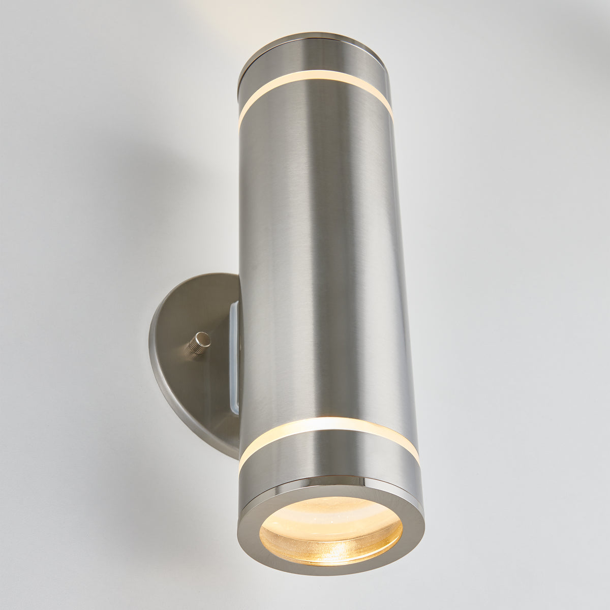 A modern, cylindrical brushed metal wall sconce emits light from both the top and bottom, mounted on an oval metal backplate against a white wall.