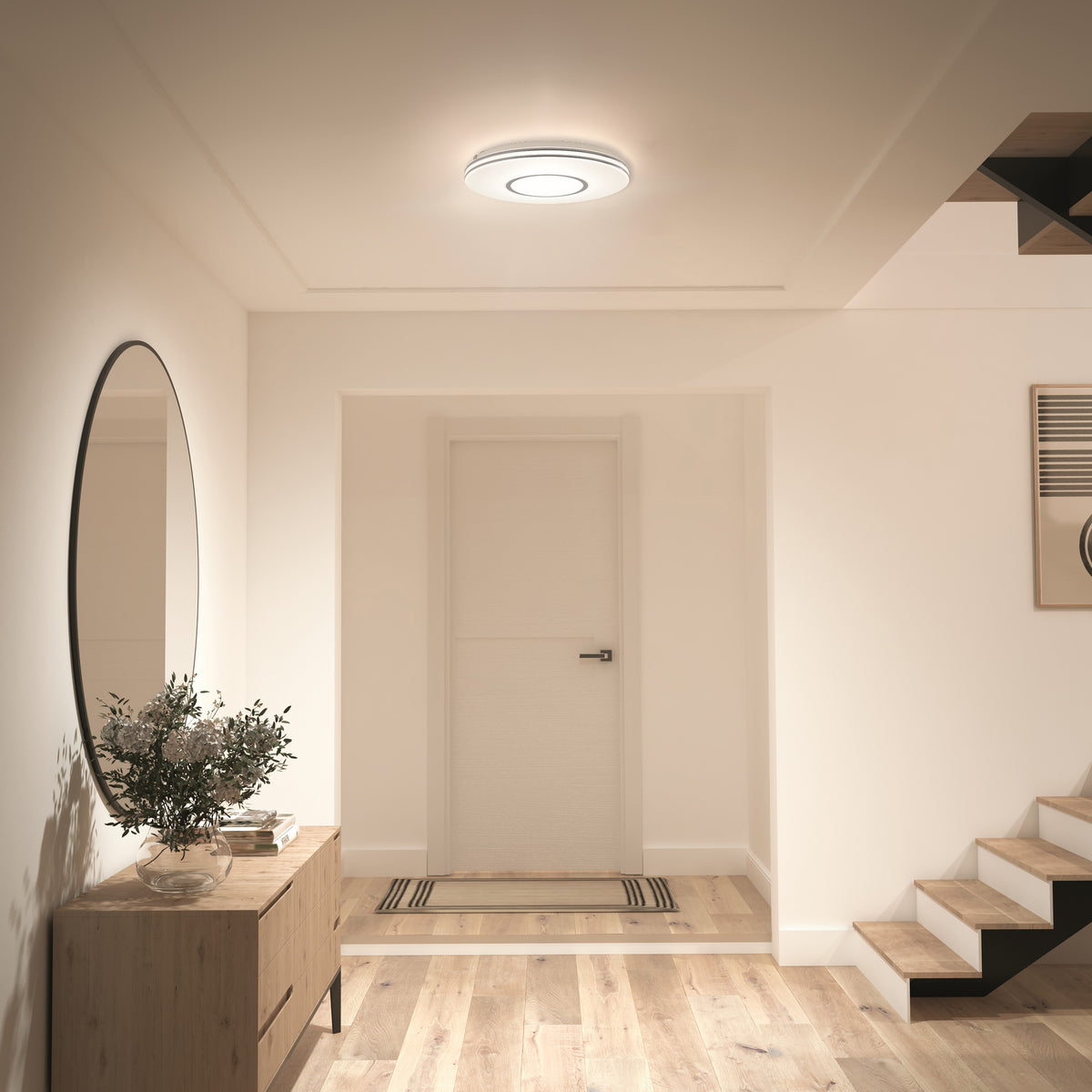 A modern hallway with light wood floors, a round mirror above a wooden console table with a plant, a white door at the end, and an illuminated ceiling light. A staircase with wooden steps is on the right.