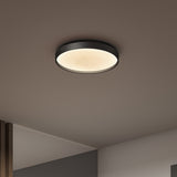 A round, flush-mounted ceiling light with a black frame and diffused white glow is illuminated on a light-colored ceiling in a modern interior space.
