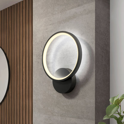 The Artika Solis modern LED indoor-outdoor wall light 5 CCT, featuring a glowing circular design, is mounted on a gray wall with vertical wooden slats and a round mirror to the left and green plant leaves in the bottom right corner.