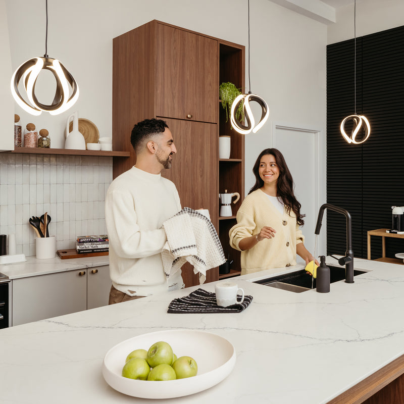 A man and woman smile while drying dishes together in a modern kitchen with wooden cabinets, a bowl of green apples on the counter, and an Artika Vivaldi LED pendant light 3 CCT overhead.