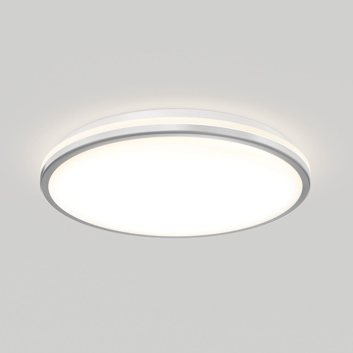 A modern, round, flush-mounted ceiling light with a white diffuser and a slim metallic rim, emitting a soft, even glow against a plain light gray ceiling.
