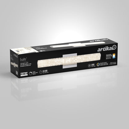 A rectangular black Artika box displays the Exalto LED bathroom vanity light, featuring a crystal-accented fixture image, product details, and icons for brightness, lifespan, and energy efficiency.