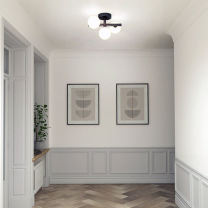 A hallway with light gray paneled walls, two abstract art prints in frames, a wooden herringbone floor, a potted plant on the window sill, and Artikas Bloom LED flush mount ceiling light with three round bulbs.