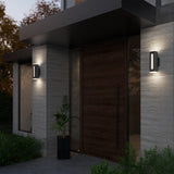 A modern house entrance with a large wooden door, two wall-mounted rectangular lights on either side, a black planter with a green plant, and surrounding greenery at dusk.