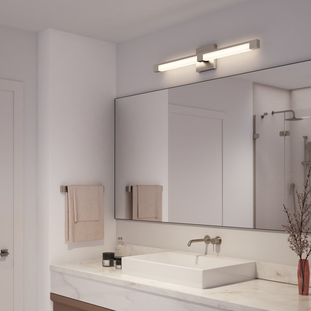 Modern bathroom featuring the Artika Tivoli modern LED bathroom vanity light (5 CCT) above a large rectangular mirror, white sink on marble countertop, beige towels, and a vase with dried branches. Shower visible in the reflection.