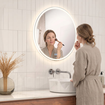 A woman in a beige robe applies makeup at a sink, looking into the illuminated Emeraude anti fog LED wall mirror by Artika on the tiled wall. A vase with dried grass is placed on the counter.