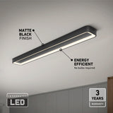 A matte black integrated LED ceiling light fixture is mounted on a white ceiling, labeled as energy efficient with no bulbs required and a 3-year warranty. The room features neutral-colored walls and cabinets.