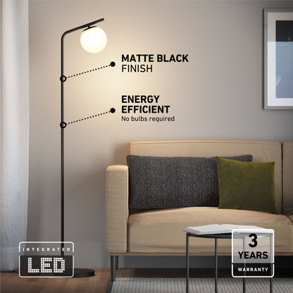 The Artika Clayton dimmable LED modern mid-century floor lamp, with a matte black finish and energy-efficient round LED light, stands by a beige sofa with dark and green cushions. Includes a “3 Years Warranty” label.