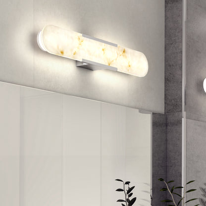 The Artika Selena 3 CCT LED bathroom vanity light features a frosted, elongated glass shade and emits a soft, warm glow above a bathroom mirror on a gray tiled wall, with plant leaves in the foreground.