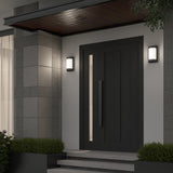 Modern front door entrance with dark wood door, vertical window panel, sleek handle, gray tiled walls, two lit wall sconces, and small shrubs on both sides of the steps.