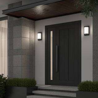 A modern front door with a vertical handle is flanked by two Artika Fogo modern LED indoor-outdoor wall lights, set against gray tiled walls with concrete steps and green shrubs on either side.