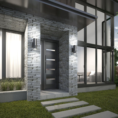 Modern house entrance with a gray stone exterior, sleek black door with horizontal glass panels, and two Artika Linea modern indoor-outdoor LED wall lights beside large windows showing a living room sofa. Concrete stepping stones lead to the door.