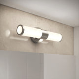 A modern wall-mounted light fixture with a cylindrical frosted glass shade and metallic base, installed on a light-colored tiled wall above a mirror. The fixture emits soft, even lighting.