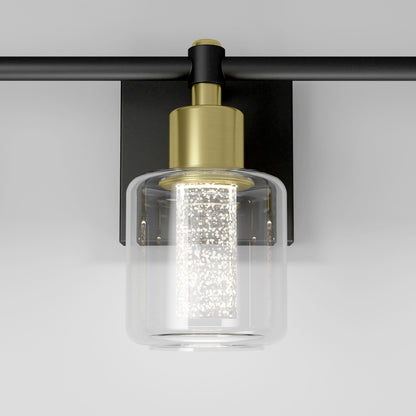 The Artika Champagne Globe bathroom vanity light features a black mount, brass socket, and clear cylindrical glass shade with a textured illuminated bulb, making it a modern accent against any light background.