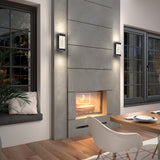 Modern living room with a gray concrete fireplace, a fire burning inside, wall lights on either side, large windows, a wooden dining table set, and a cushioned window seat with a plant.