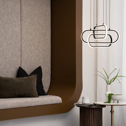 A cozy alcove features a cushioned bench with black and brown pillows, a round side table holding a plant, mug, and book, and is illuminated by the Artika Lewis modern LED pendant light 3 CCT against a brown and white backdrop.