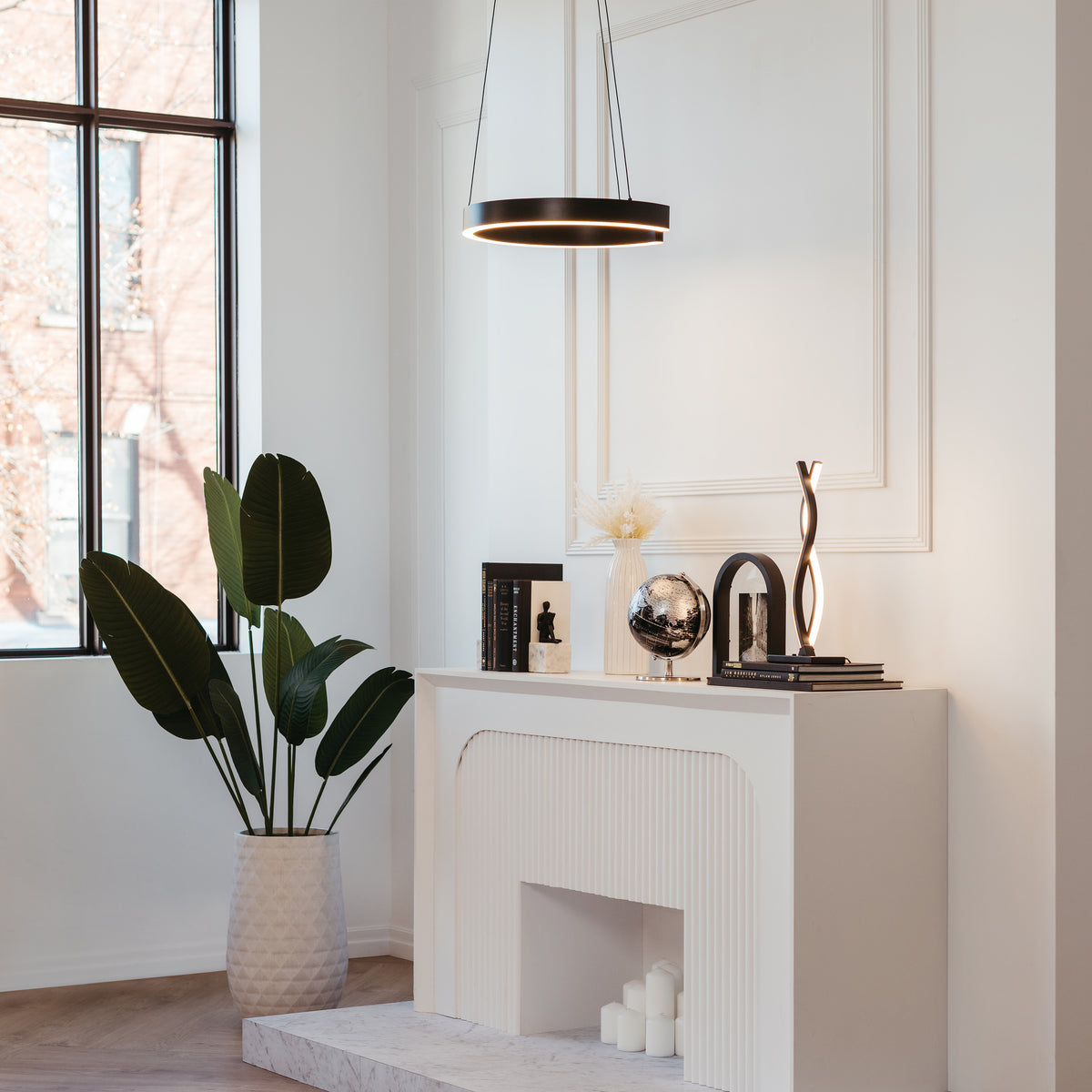 A modern, minimalist living room features a white fireplace with decorative items, a large potted plant, and a round pendant light hanging above. Tall windows let in natural light, creating a bright, airy atmosphere.