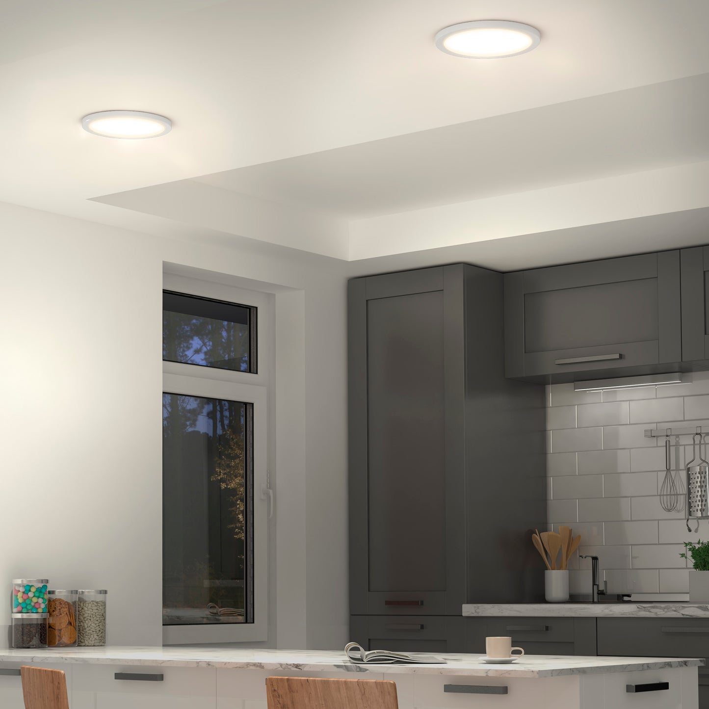 Modern kitchen featuring gray cabinets, white countertops, a white tile backsplash, visible utensils, an outdoor-view window, and Artikas Europa Disk 9 in. LED modern flush mount light (3 CCT) illuminating the space.