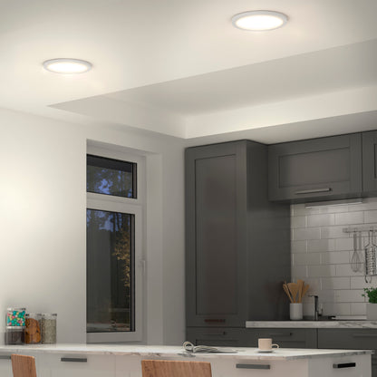 Modern kitchen featuring gray cabinets, white countertops, a white tile backsplash, visible utensils, an outdoor-view window, and Artikas Europa Disk 9 in. LED modern flush mount light (3 CCT) illuminating the space.