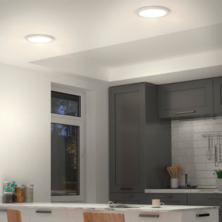 Modern kitchen with gray cabinets, white tile backsplash, and a window with a view of trees. Two Artika Europa Disk 9 in. LED modern flush mount lights brighten the space. A white island with chairs and kitchenware is in front.