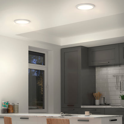 Modern kitchen with gray cabinets, white tile backsplash, and a window with a view of trees. Two Artika Europa Disk 9 in. LED modern flush mount lights brighten the space. A white island with chairs and kitchenware is in front.
