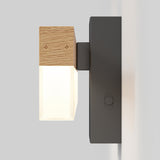 A modern wall-mounted light fixture with a black rectangular base, a wooden top section featuring plus and minus symbols, and a frosted glass shade emitting a soft light.