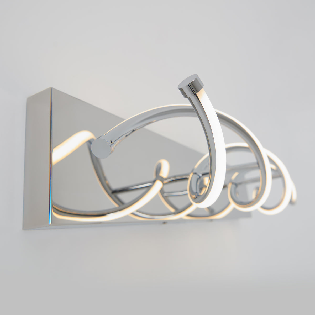 A modern wall-mounted LED light fixture with chrome finish and looped, curved design, emitting a soft white glow against a light gray wall.