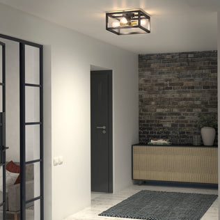 A modern hallway features the Artika Princeton 4-light flush mount ceiling light with a black frame, gray door, exposed brick accent wall, console table with books and a plant, glass-paneled door, and a dark textured rug on a light floor.
