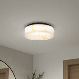 A modern, round ceiling light with a white and beige marbled pattern is mounted on a white ceiling. Part of a mirror, plant, and wooden door frames are visible in the minimalist room below.