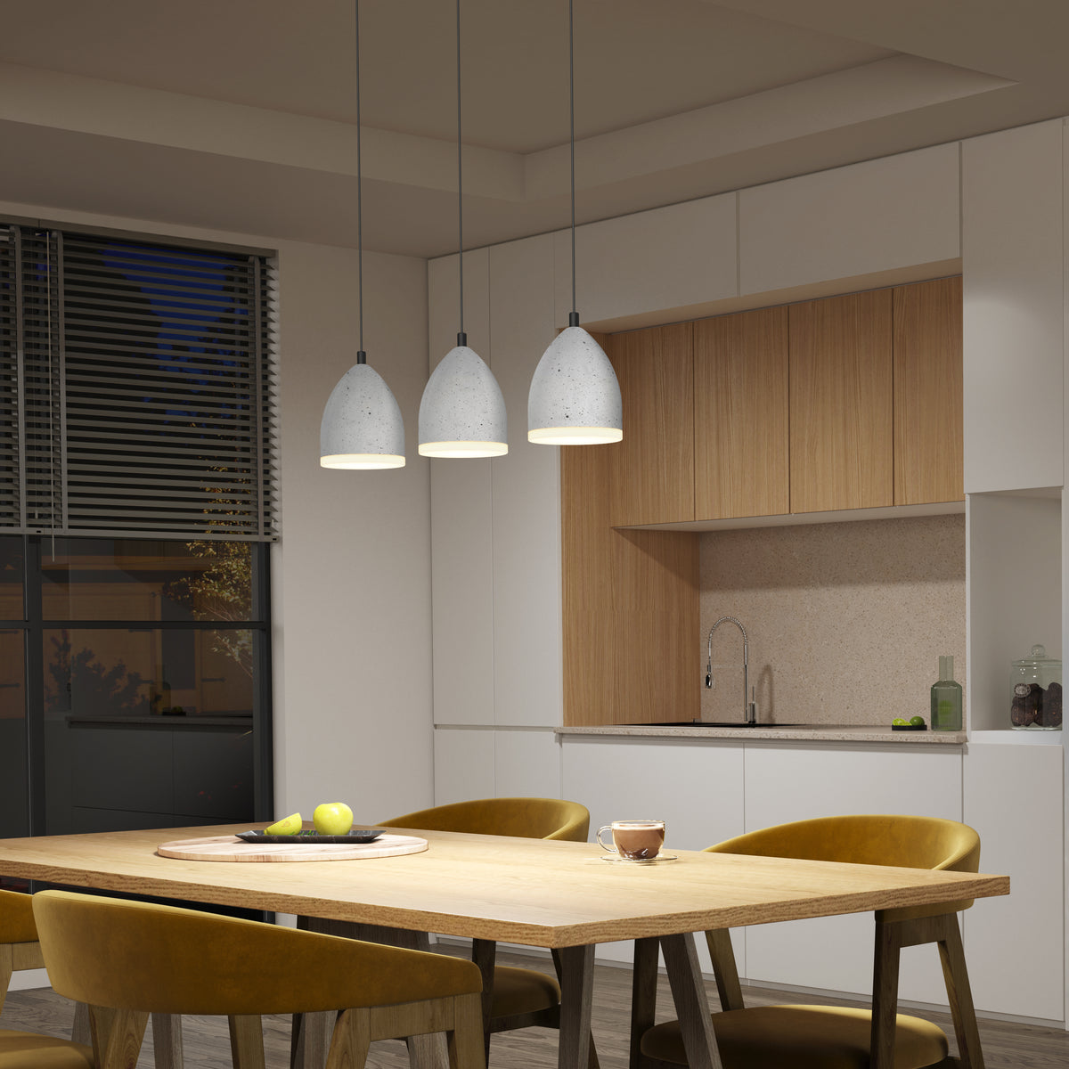 A modern kitchen with white and wood cabinets, a wooden dining table, yellow chairs, three hanging pendant lights, and a window with blinds. A bowl of apples and a mug are on the table.