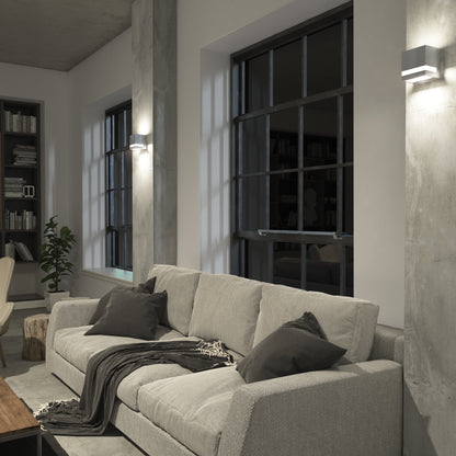 A modern living room with a light gray sofa, dark pillows, and a throw blanket by large windows and concrete columns features Artikas Valor Square LED indoor-outdoor wall lights and a bookshelf filled with books.
