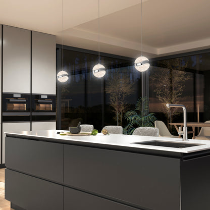 Modern kitchen with a large island, built-in ovens, a sink, and three Artika Wavey LED pendant lights. Floor-to-ceiling windows reveal trees outside, with a dining area visible in the background.