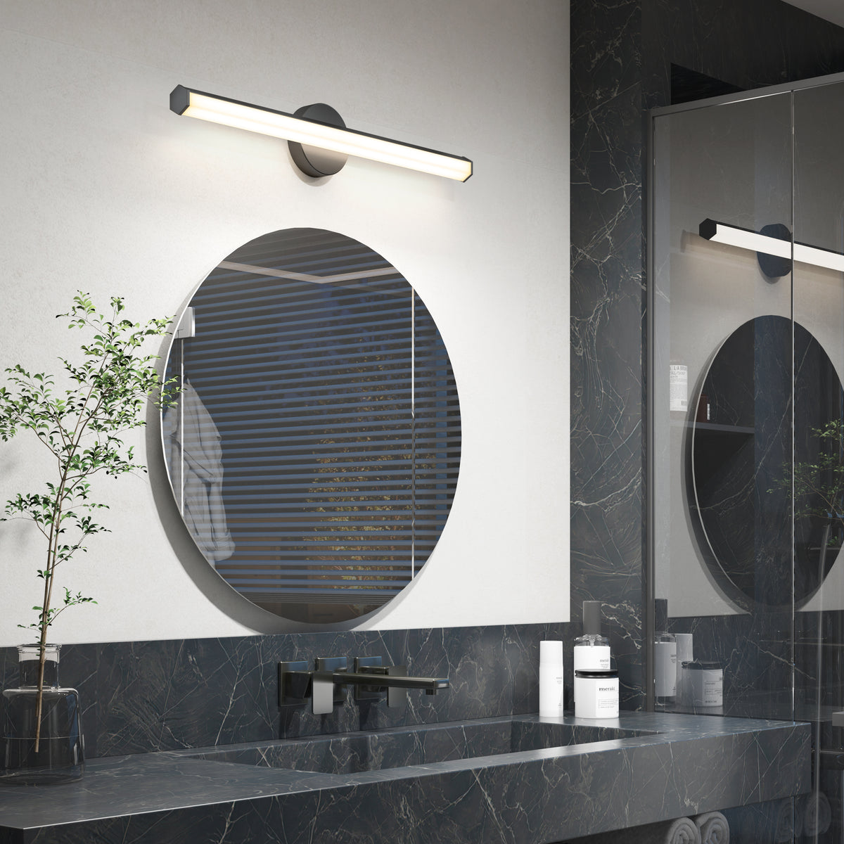 Modern bathroom with a round mirror, sleek black sink, and countertop with toiletries. A wall-mounted light fixture is above the mirror. There is a small plant on the counter and a reflection of blinds in the mirror.