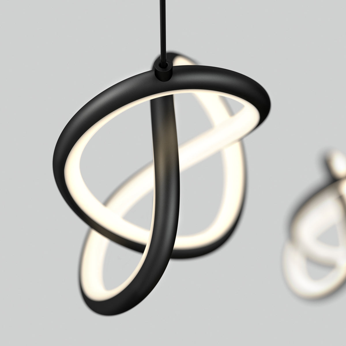A modern, sculptural pendant light with two interlocking, curved black rings emitting a soft white glow, suspended against a plain light gray background. A blurred similar light is visible in the background.