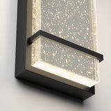Close-up of a modern wall sconce featuring a rectangular glass panel with tiny bubbles inside, illuminated by soft white lights, and framed with sleek black metal.