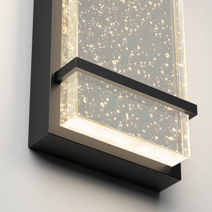 The Artika Skyler indoor-outdoor LED wall light features a rectangular black frame and a clear, bubble-filled glass panel that emits a warm glow when illuminated.