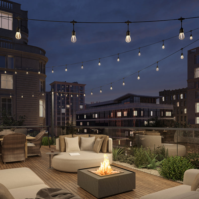 A cozy rooftop patio at dusk is illuminated by Artika's Eclipse Solar String Light with Remote, with modern outdoor furniture, a blazing fire pit, and city buildings under a darkening sky.