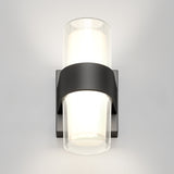 A modern wall sconce with a black rectangular base and two cylindrical glass shades, emitting soft white light, mounted on a plain light-colored wall.