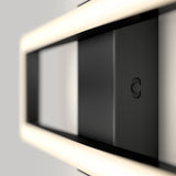 A close-up view of a modern, rectangular black light fixture with soft white LED lighting along its edges, mounted on a light gray wall. The design is minimalistic and geometric.