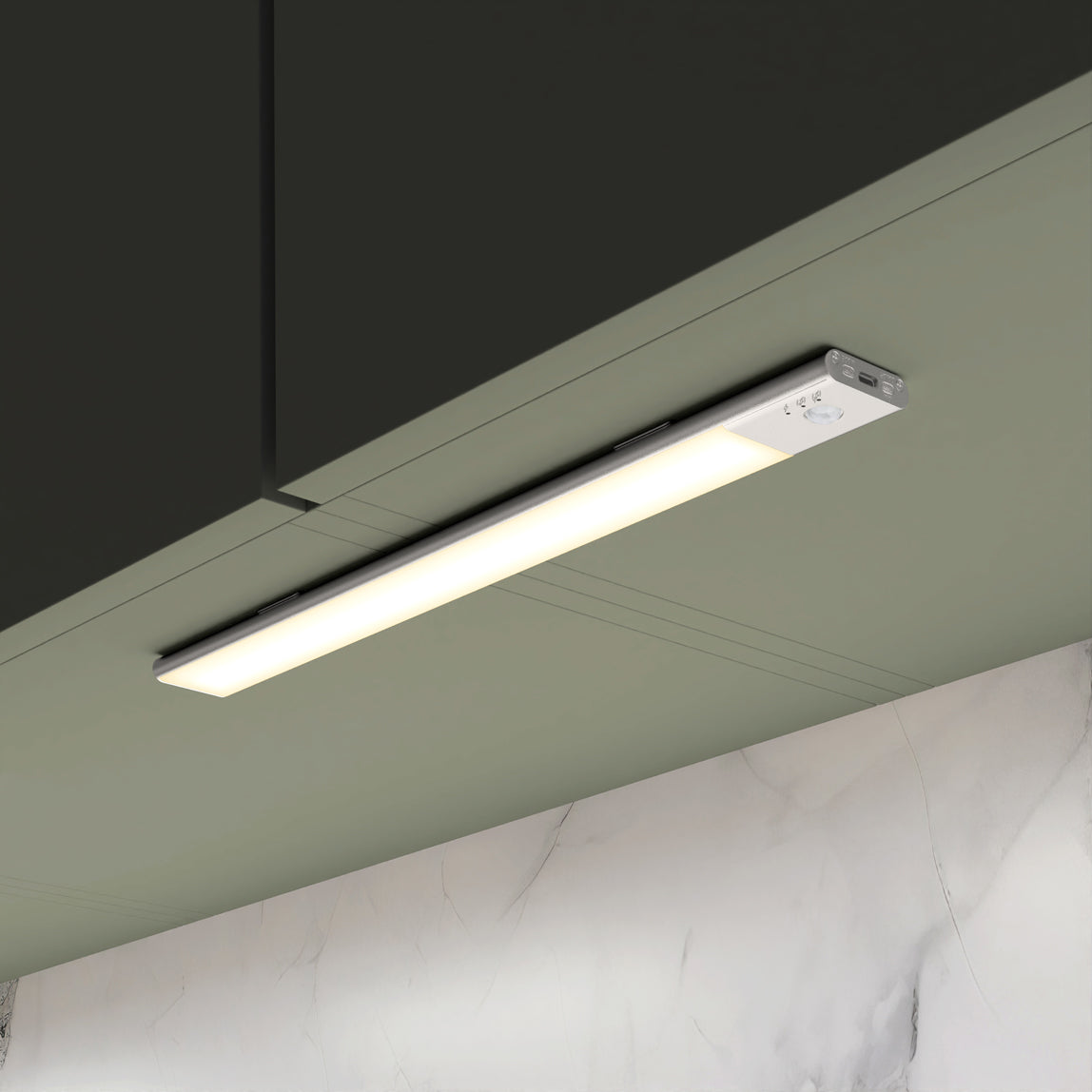 A slim, rectangular LED light fixture is mounted under a dark green cabinet, illuminating a light green wall and a marble backsplash below.