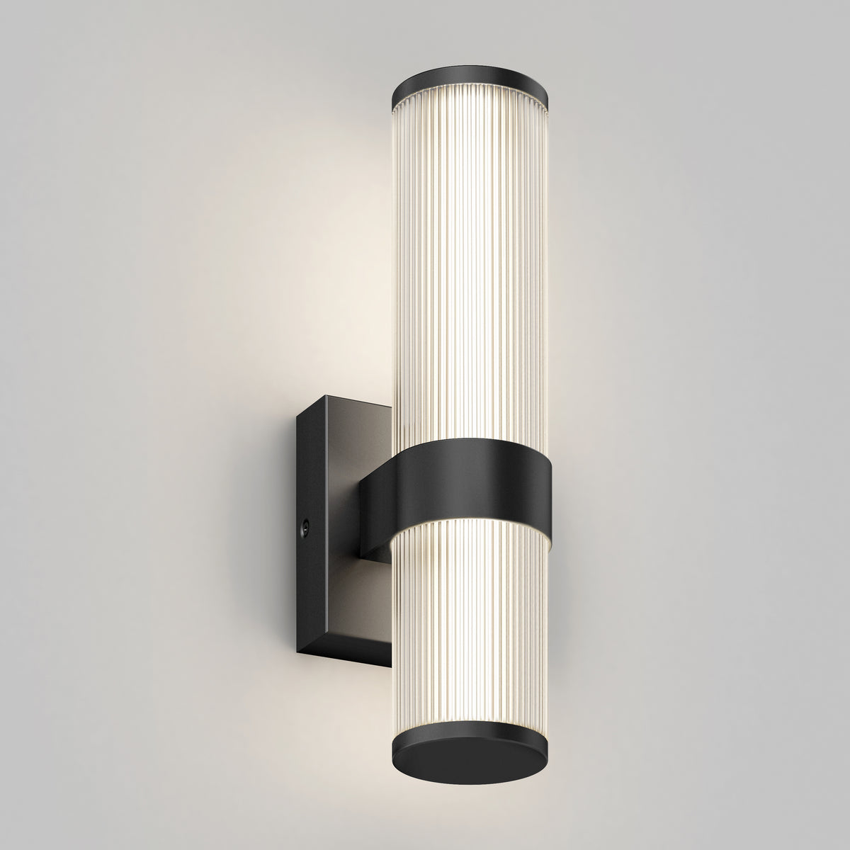 A modern wall sconce with a vertically oriented, ribbed cylindrical glass shade and black metal accents mounted on a square wall plate. The light emits a soft, diffused glow.