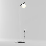 A modern, minimalist black floor lamp with a round white bulb at the top stands near a white wall outlet. The lamp is plugged in, and the cord is visible running to the socket. The background is plain and light-colored.
