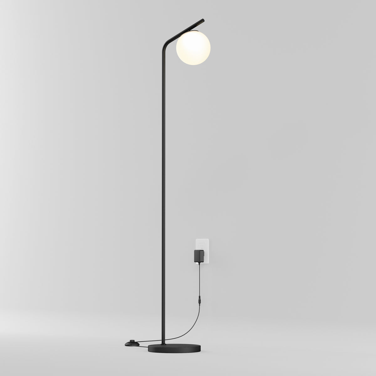 The Artika Clayton dimmable LED modern mid-century floor lamp features a minimalist black design with a round white bulb, set against a plain light gray wall. Its visible cord runs down to the base and is plugged into a nearby outlet.