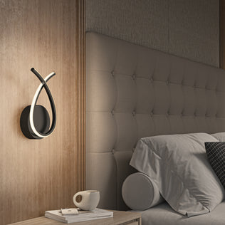 A modern bedroom features a tufted headboard, neatly made bed with gray bedding and a bolster pillow, a nightstand with book and cup, and the Artika Gamma modern LED wall sconce 5 CCT mounted on a wooden panel.