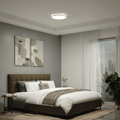 Modern bedroom with a large bed, neutral bedding, two abstract paintings on a gray wall, a potted plant by the window with sheer curtains, and an Artika Hemisphere 15 in. LED flush mount light (5 CCT) brightening the space.