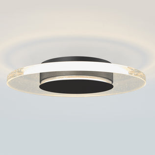 The Artika Essence Disk LED flush mount features a modern circular design with a black central base and a glowing, translucent outer ring accented by sparkling details, casting a soft, diffused glow onto the ceiling.